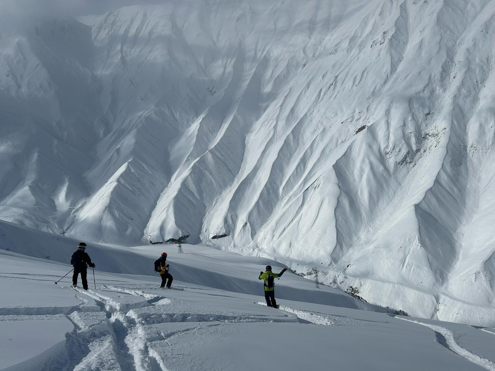 Skiers and snowboarders in deep powder look across to a steep, fluted snow face in Gudauri