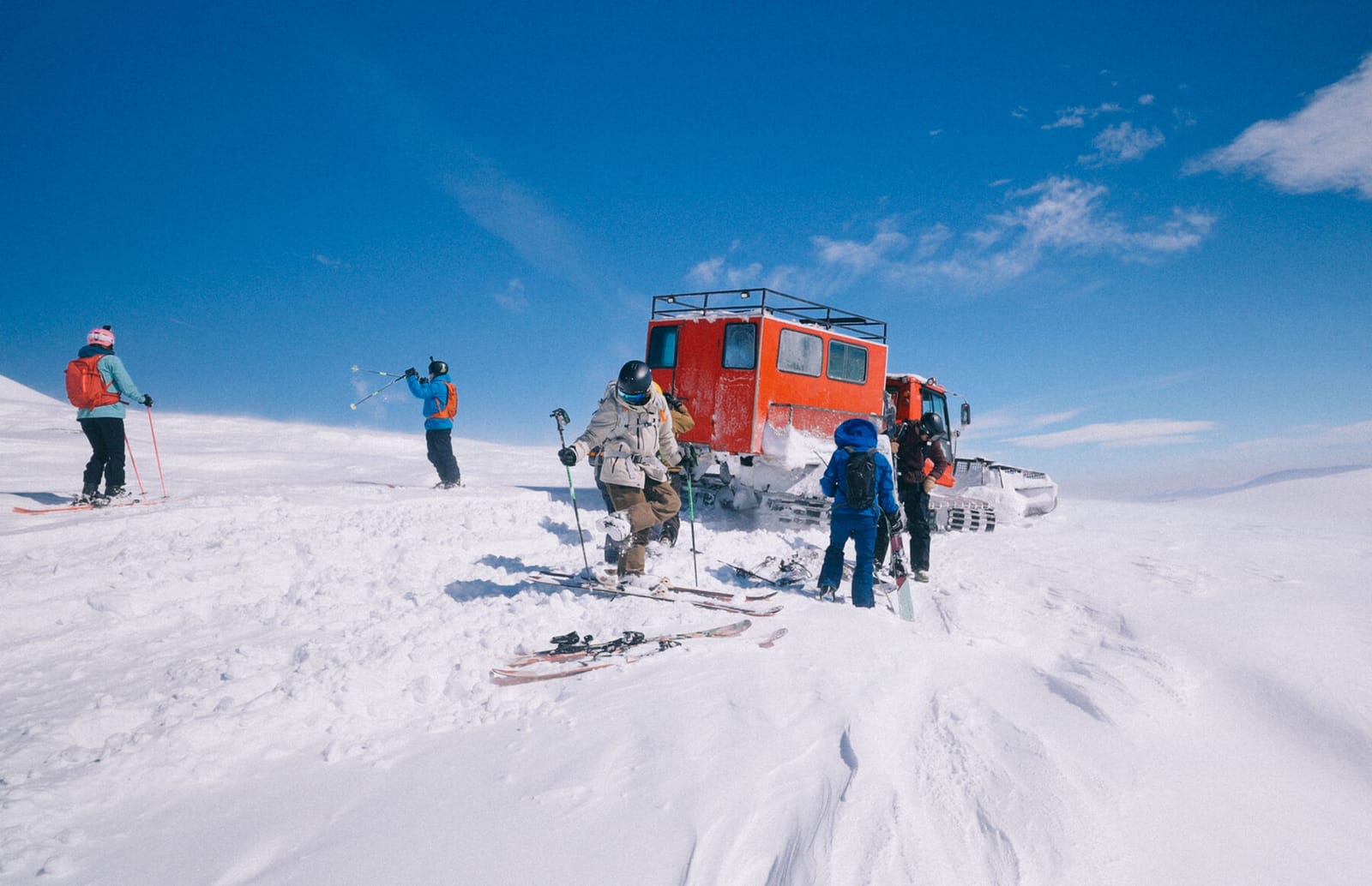 Riders clipping in beside an orange snowcat under a blue sky in Goderdzi