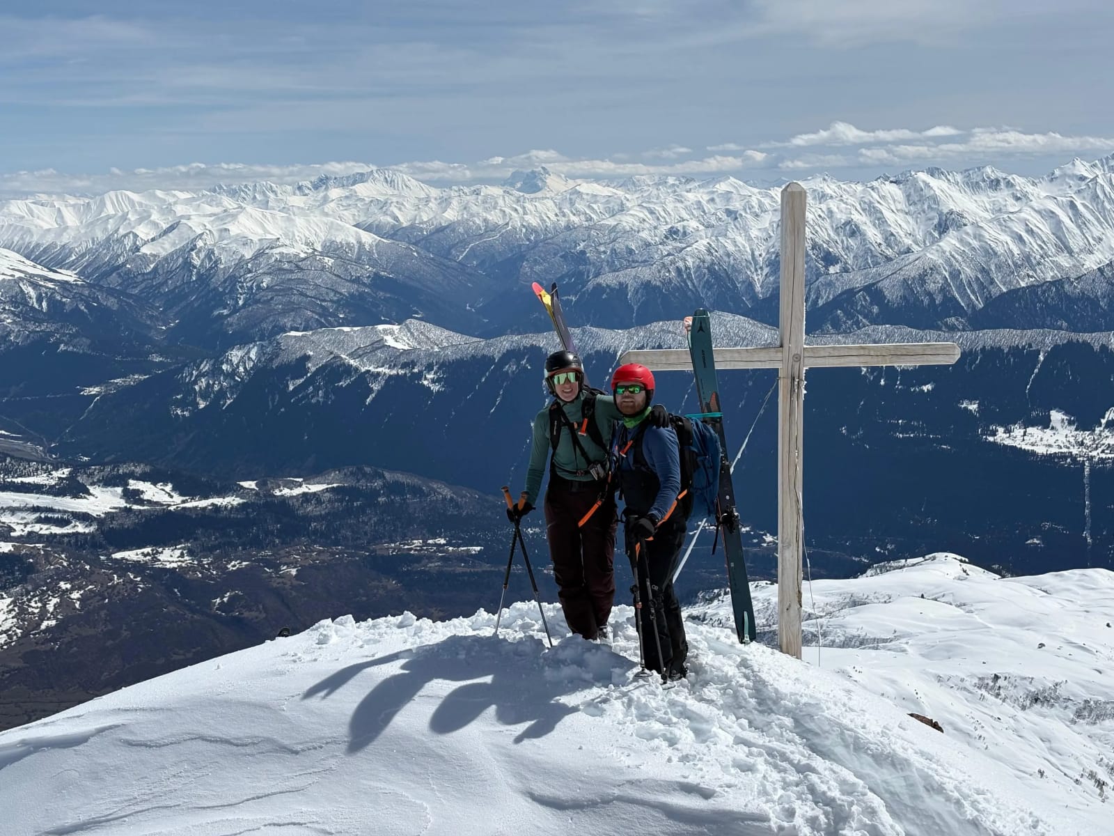 Two skiers beside a wooden summit cross above Gudauri mountain valleys