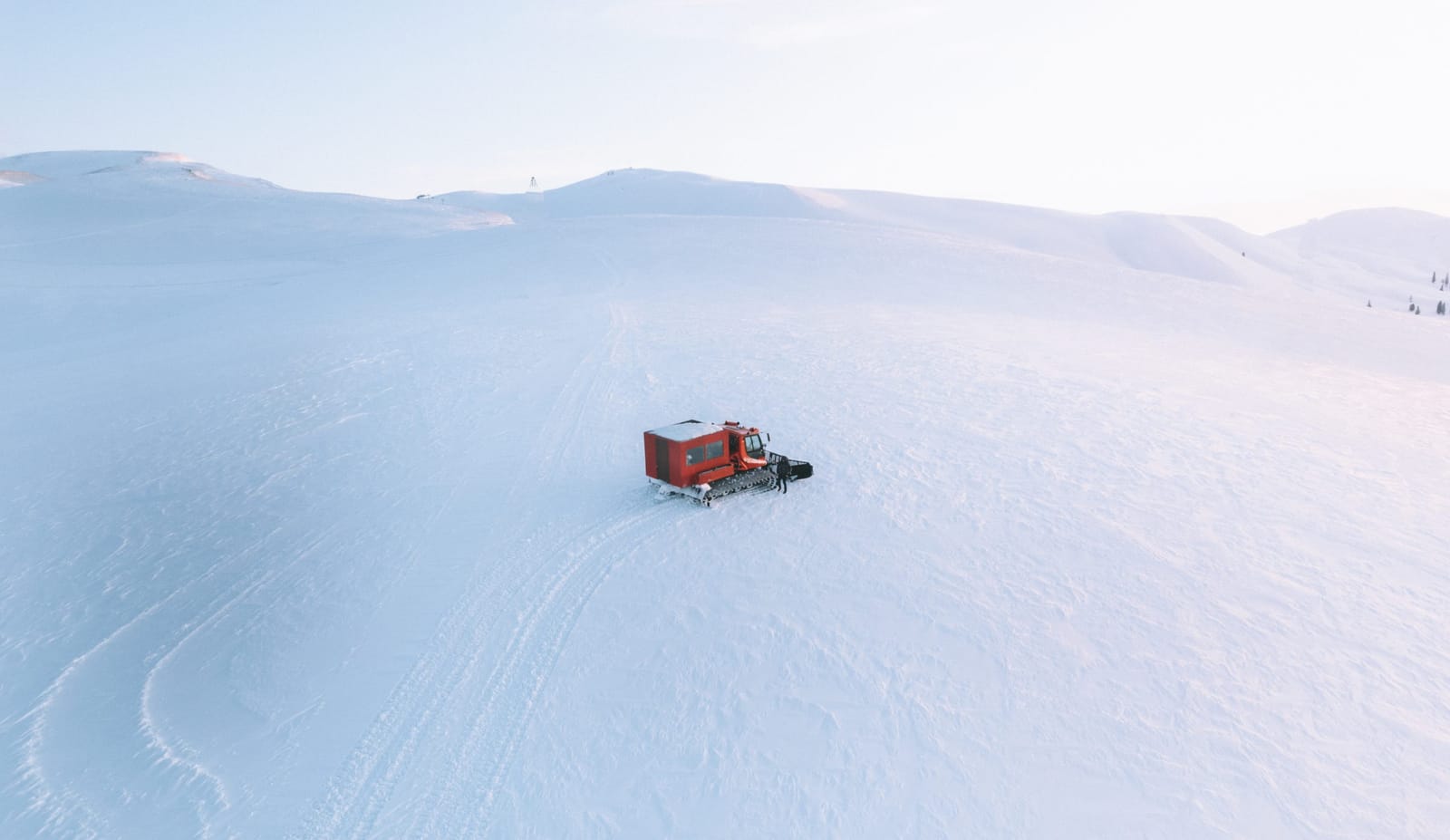 An orange snowcat on a wide, snow-covered slope with ski tracks, seen from above