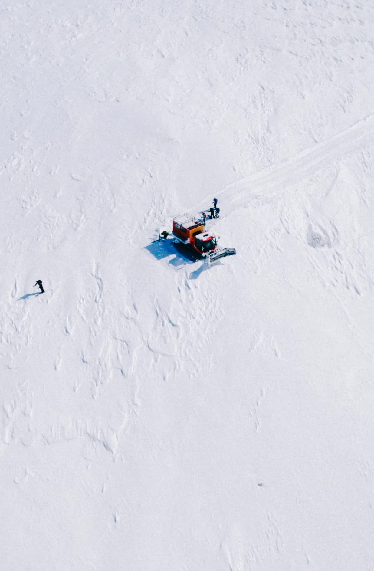 Aerial view of a red snowcat on a wide white snow slope with a few riders standing beside it and one skier below