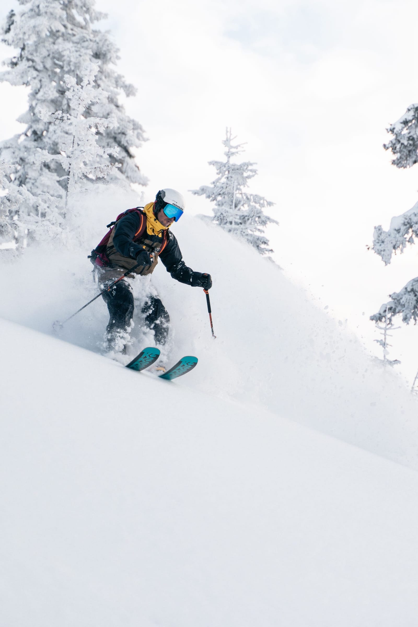 A skier in a yellow jacket and blue goggles riding powder beside snow-covered fir trees in Svaneti