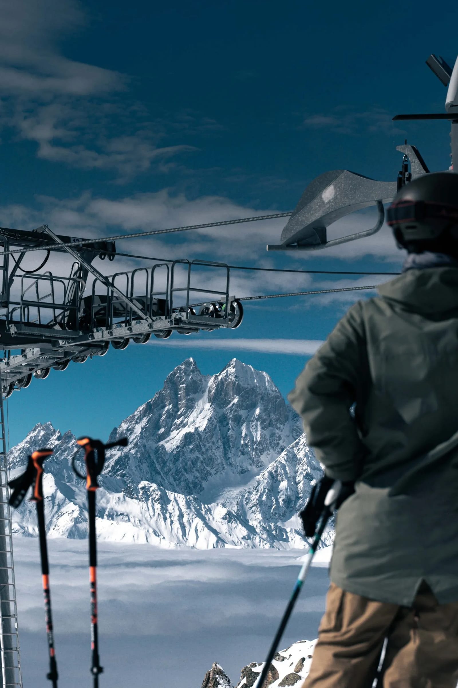 A skier in an olive jacket seen from behind at a Tetnuldi lift station, ski poles in the foreground and the twin peaks of Ushba above a sea of cloud