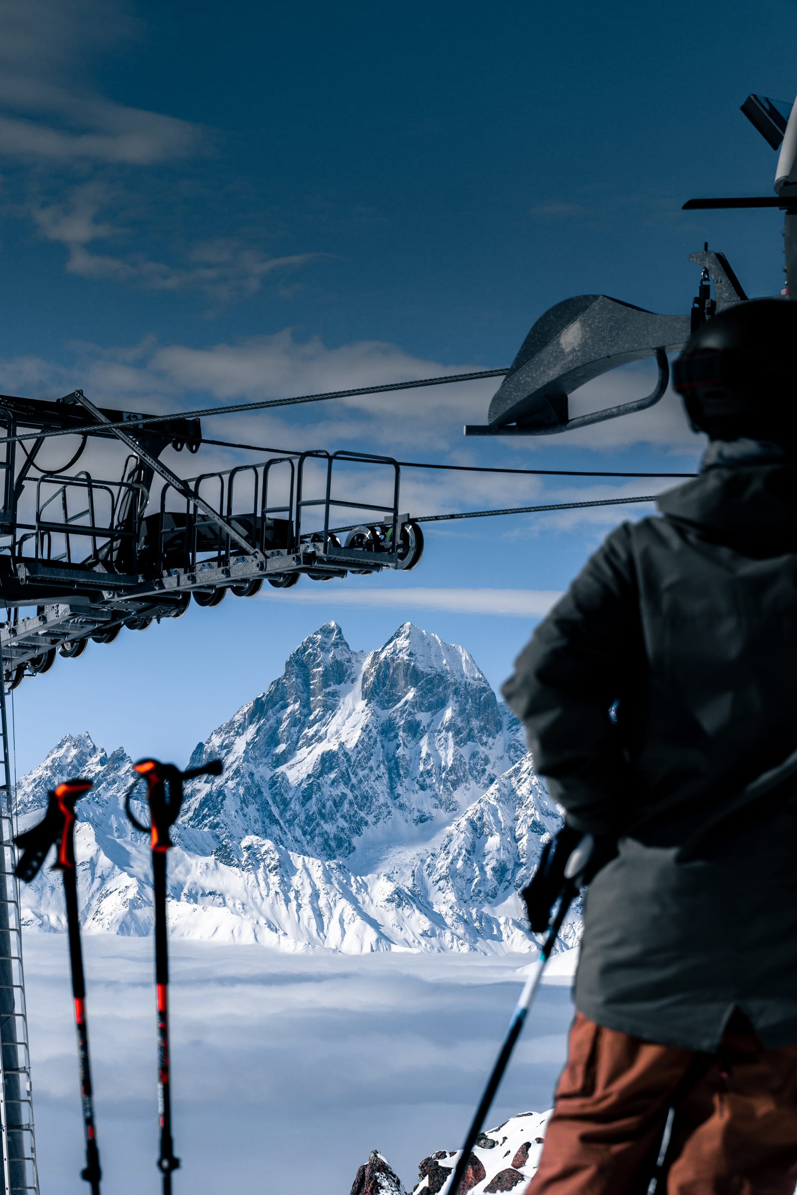 A skier seen from behind at a lift station, with ski poles in the foreground, looks across a sea of cloud at Ushba, the twin-peaked mountain in Svaneti